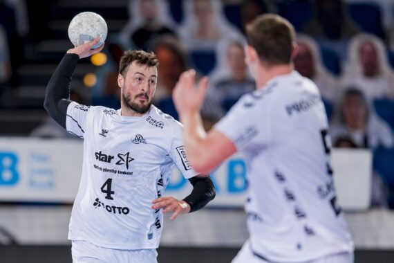 Ein männlicher Handballspieler in einem weißen Trikot mit der Nummer 4 bereitet sich auf den Wurf vor, während ein anderer Spieler in einem weißen Trikot im Vordergrund steht und ihm teilweise gegenübersteht. Beide befinden sich auf einem Hallenplatz mit unscharfen Zuschauern im Hintergrund.