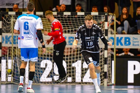 Ein Handballspieler in einem schwarzen Trikot jubelt beim Laufen, während ein anderer Spieler in einem weiß-blauen Trikot in der Nähe steht. Im Hintergrund sind ein Torwart und Zuschauer zu sehen.