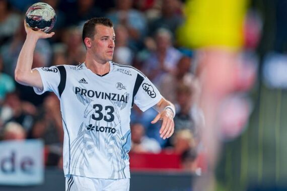 Ein männlicher Handballspieler in einem weißen Trikot mit der Nummer 33 bereitet sich darauf vor, den Ball während eines Spiels zu werfen. Die Zuschauer sind im Hintergrund verschwommen, und eine gelb gekleidete Figur ist teilweise im Vordergrund zu sehen.