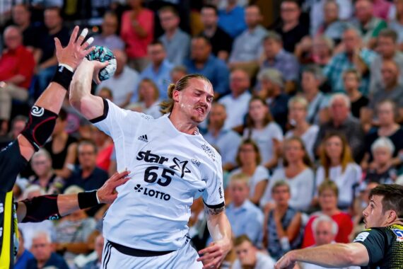 Ein Handballspieler im weißen Trikot mit der Nummer 65 springt in der Luft, um den Ball zu werfen, während die Verteidiger in schwarzen und gelben Trikots versuchen, ihn zu blockieren. Eine Zuschauermenge beobachtet das intensive Geschehen.