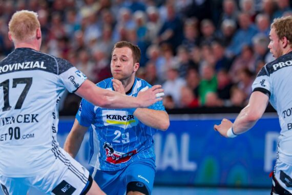 Ein Handballspieler in einem blauen Trikot läuft mit dem Ball nach vorne und sieht sich zwei Verteidigern in weißen Trikots auf einem Spielfeld mit einem unscharfen Publikum im Hintergrund gegenüber.