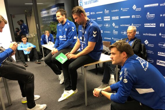 Eine Gruppe von Männern in blauer Sportkleidung sitzt und steht um Tische in einem Konferenzraum, hinter dem eine blaue Wand mit Sponsorenlogos hängt. Einige schreiben, während andere aufmerksam zuhören.