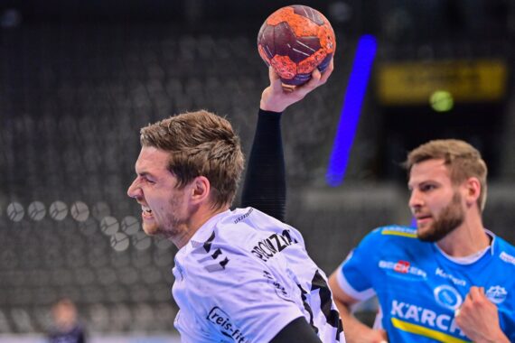 Ein männlicher Handballspieler in einem weißen Trikot bereitet sich mit konzentrierter Miene auf den Wurf vor, während ein Verteidiger in einem blau-gelben Trikot in einer Sporthalle in der Nähe steht.
