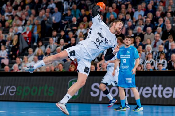 Ein männlicher Handballspieler in einem weißen Trikot springt in die Luft, um den Ball zu werfen, während im Hintergrund Zuschauer und ein anderer Spieler in Blau zusehen.