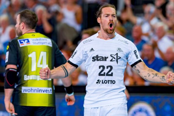 Ein Handballspieler im weißen Trikot mit der Nummer 22 jubelt leidenschaftlich auf dem Spielfeld, ballt die Fäuste und schreit, während ein anderer Spieler im schwarz-gelben Trikot mit der Nummer 14 im Hintergrund davonläuft.