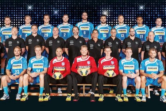 Eine Herren-Handballmannschaft posiert für ein Gruppenfoto in blau-weißen Trikots, wobei drei Personen in roten Trikots Bälle halten. Die schwarz gekleideten Trainer stehen auf beiden Seiten vor einem gemusterten, beleuchteten Hintergrund.