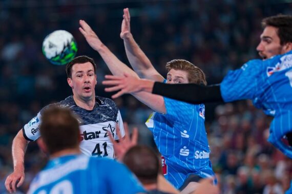 Ein Handballspieler in einem dunklen Trikot bereitet sich darauf vor, den Ball zu werfen, während drei Verteidiger in blauen Trikots ihre Arme heben, um ihn zu blockieren, und alle sind während eines Spiels sehr konzentriert.
