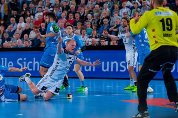 Ein Handballspieler in einem weißen Trikot stürzt, während er den Ball in Richtung Tor wirft, umgeben von Spielern in blauen Trikots, während sich ein Torwart in Gelb darauf vorbereitet, den Schuss zu blocken. Eine Menschenmenge schaut im Hintergrund zu.