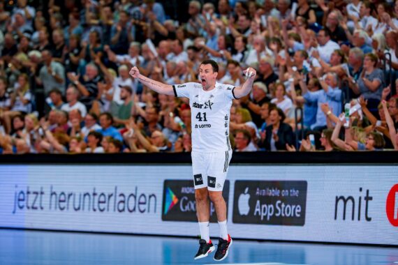 Ein Handballspieler in einem weißen Trikot jubelt mit erhobenen Armen vor einer jubelnden Menge in einer vollen Arena. Das Publikum steht und klatscht und schafft eine energiegeladene Atmosphäre.
