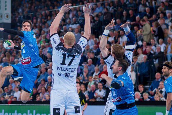 Ein Handballspieler in Blau springt, um den Ball zu schießen, während drei Verteidiger in Weiß aufspringen, um ihn zu blockieren.