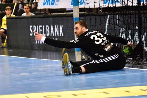 Ein Handballtorwart in einem schwarzen Trikot mit der Nummer 33 auf dem Rücken sitzt während eines Spiels auf dem Boden in der Nähe des Torpfostens und schaut nach links, während im Hintergrund Zuschauer zu sehen sind.