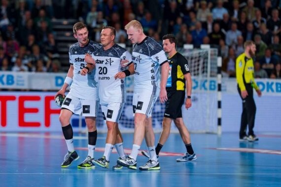 Drei männliche Handballspieler in weißen Trikots helfen einem verletzten Mannschaftskameraden vom Spielfeld, während im Hintergrund ein Schiedsrichter und ein Torwart stehen. Die Szene findet in einer Halle statt, in der eine Menschenmenge zuschaut.
