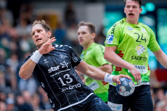 Ein Handballspieler in einem schwarzen Trikot bereitet sich darauf vor, den Ball zu werfen, während er während eines Spiels von zwei Spielern in grünen Trikots eng verteidigt wird. Die Szene ist intensiv, die Zuschauer sind im Hintergrund verschwommen.