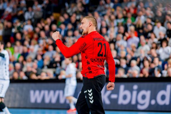 Ein Handballspieler in einem roten Trikot mit der Nummer 21 ballt auf dem Spielfeld jubelnd die Faust, während im Hintergrund unscharfe Zuschauer und Gegner in weißen Trikots zu sehen sind.