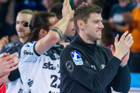Eine Gruppe von männlichen Sportlern in Sportkleidung klatscht und feiert, wobei ein Mann im Vordergrund lächelt und andere im Hintergrund klatschen, einige mit erhobenen Händen.
