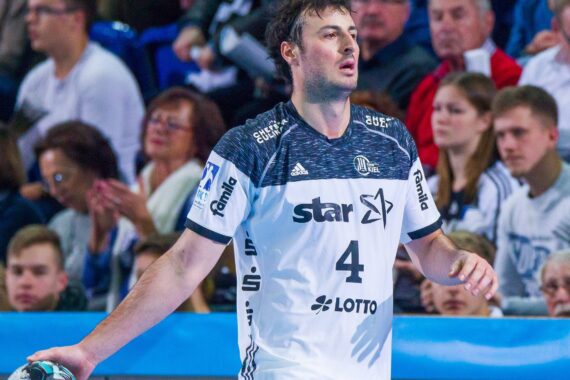 Ein männlicher Handballspieler in einem weiß-marinefarbenen Trikot mit der Nummer 4 steht auf dem Spielfeld und hält einen Ball. Im Hintergrund sitzen Zuschauer und schauen zu.