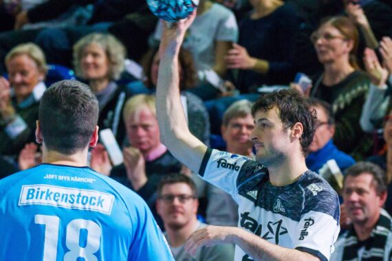Ein Handballspieler in einem weißen Trikot bereitet sich darauf vor, den Ball zu werfen, während er einem Gegner in einem blauen Trikot mit der Nummer 18 gegenübersteht, während das Publikum im Hintergrund zusieht und applaudiert.