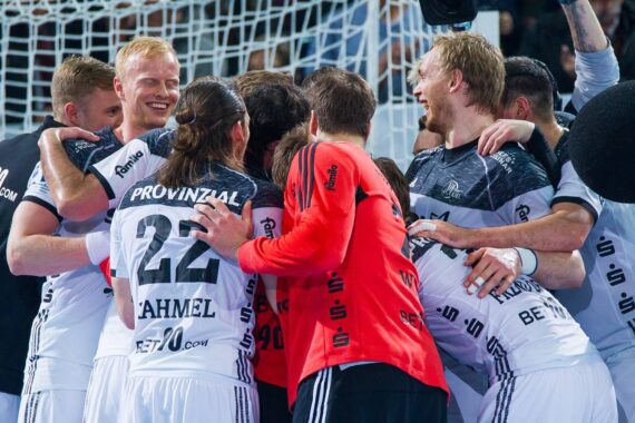Eine Gruppe von Handballspielern in weißen und schwarzen Trikots drängt sich jubelnd, lächelnd und einander umarmend auf dem Spielfeld vor einem Netz zusammen.