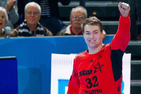 Ein Handballspieler in einem roten Trikot mit der Nummer 32 hebt die Faust und lächelt, während er vor einem Publikum in einer Sportarena steht.