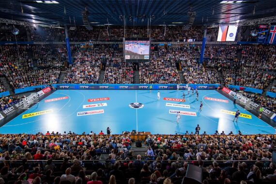 Eine große Sporthalle, gefüllt mit Zuschauern, die ein Handballspiel auf einem hellblauen Spielfeld verfolgen, mit Spielern in Aktion und einer Anzeigetafel, die über der Mitte hängt.