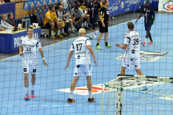 Drei Handballspieler in weißen Trikots stehen während eines Spiels auf einem blauen Hallenplatz, mit einem Netz im Vordergrund und Zuschauern, die im Hintergrund sitzen.