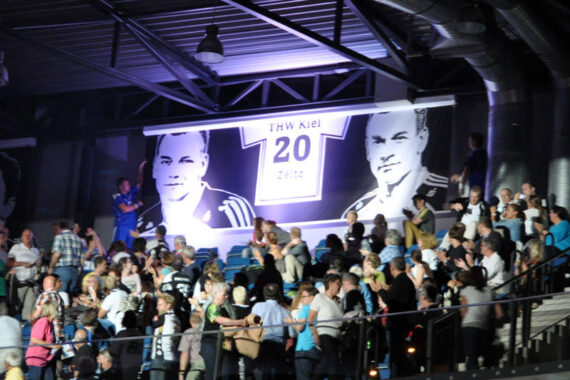 Eine große Menschenmenge versammelt sich in einer Halle unter einem Banner mit einem Trikot des THW Kiel 20 Zeitz und dem Porträt eines Mannes, während Scheinwerfer die Szene beleuchten.
