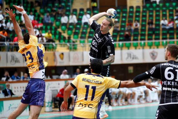 Ein Handballspieler in einem schwarzen Trikot springt und wirft den Ball, während zwei Spieler in Gelb versuchen, ihn während eines Hallenspiels zu blockieren. Die Zuschauer sind im Hintergrund auf grünen Sitzen zu sehen.