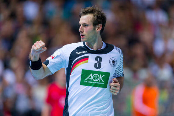 Ein männlicher Handballspieler, der ein weißes Deutschland-Trikot mit der Nummer 3 trägt, ballt während eines Spiels die Faust, während die Zuschauer und Spieler im Hintergrund verschwommen zu sehen sind.