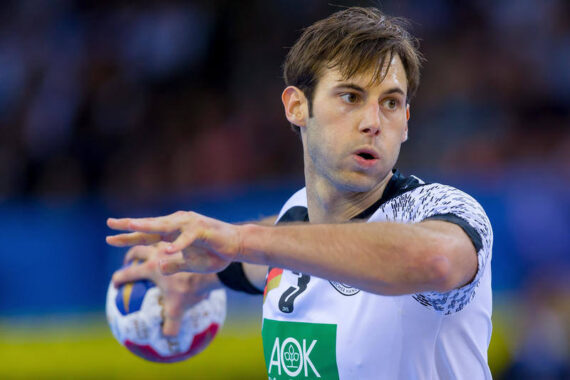 Ein männlicher Handballspieler in einem weiß-schwarzen Trikot bereitet sich darauf vor, den Ball während eines Spiels zu werfen. Er sieht konzentriert aus, seine Hände sind bereit, den Ball loszulassen. Der Hintergrund ist unscharf mit Zuschauern.
