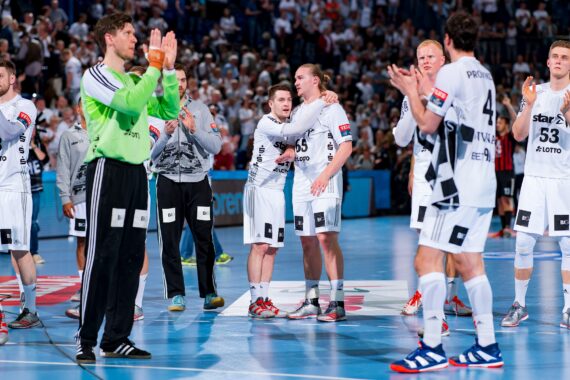 Eine Herren-Handballmannschaft in weißen Trikots steht auf dem Spielfeld und applaudiert, in der Mitte umarmen sich zwei Spieler. Der Hintergrund zeigt eine jubelnde Menge in einer Halle.
