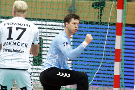Ein Handballspieler in einem hellblauen Trikot ballt die Faust und jubelt, während ein Spieler in einem weißen Trikot mit der Nummer 17 in der Nähe vor einem Netz steht.