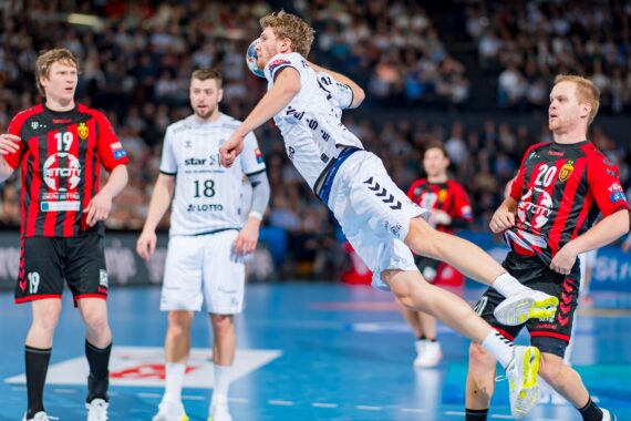 Ein Handballspieler in einem weißen Trikot springt in der Luft, um den Ball zu schießen, während Spieler in roten und schwarzen Trikots versuchen, ihn auf einem blauen Hallenplatz zu blockieren, während die Zuschauer im Hintergrund zusehen.