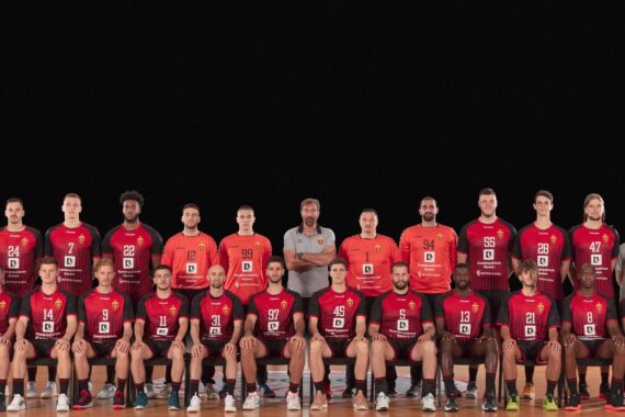 Eine Handballmannschaft stellt sich in einer Halle für ein Gruppenfoto auf. Die Spieler in kastanienbraunen Trikots sitzen und stehen in Reihen, flankiert von Trainern in grauen Hemden, alles vor einem schlichten schwarzen Hintergrund.