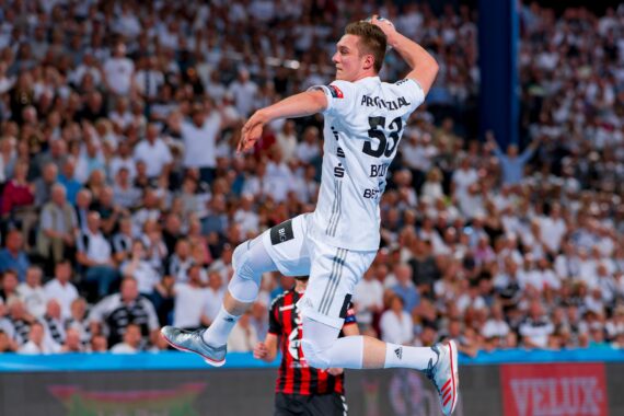 Ein männlicher Handballspieler in einem weißen Trikot springt in der Luft, um den Ball zu werfen, während im Hintergrund eine dicht gedrängte, weiß gekleidete Menschenmenge steht. Ein anderer Spieler in einer dunklen Uniform ist teilweise unter ihm zu sehen.