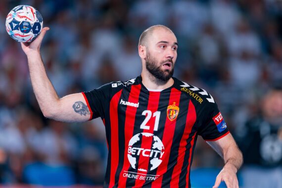 Ein männlicher Handballspieler in einem schwarz-rot gestreiften Trikot mit der Nummer 21 hält einen Ball in der rechten Hand, scheinbar bereit zum Wurf. Er ist konzentriert, mit einer unscharfen Menge im Hintergrund.