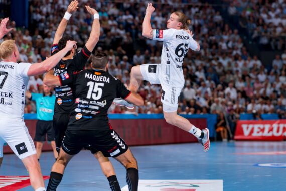 Ein Handballspieler in einem weißen Trikot springt in die Luft, um zu schießen, während drei Verteidiger in schwarzen Trikots ihre Arme heben, um ihn zu blockieren. Die Zuschauer verfolgen das Geschehen in einer voll besetzten Halle.