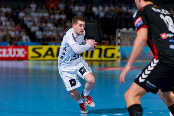 Ein Handballspieler in einem weißen Trikot läuft mit dem Ball in der Hand nach vorne, während ein Abwehrspieler in einem schwarzen Trikot sich darauf vorbereitet, ihn auf einem blauen Hallenplatz zu blocken. Die Zuschauer füllen die Tribünen im Hintergrund.