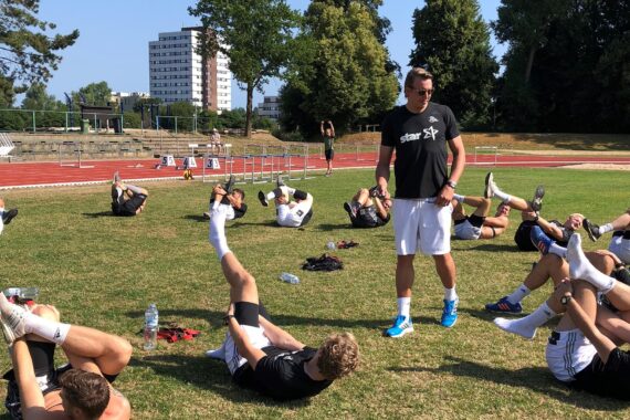 Eine Gruppe von Sportlern in Sportkleidung liegt mit angezogenen Beinen auf dem Rasen und dehnt sich auf einem Leichtathletikfeld im Freien, während ein Trainer daneben steht und beobachtet. Im Hintergrund sind Bäume und hohe Gebäude zu sehen.