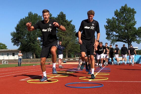 Zwei Athleten in schwarzen Hemden durchqueren beim Training im Freien auf einer roten Bahn Agility-Ringe. Im Hintergrund sind unter blauem Himmel eine Gruppe von Menschen und Bäume zu sehen.