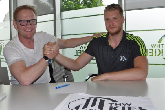Zwei Männer sitzen an einem Tisch, schütteln Hände und lächeln. Der eine trägt eine Brille und ein weißes Hemd, der andere ein schwarzes Sporthemd mit THW Kiel-Logos. Vor ihnen auf dem Tisch liegen ein Stift und eine THW-Kiel-Fahne.