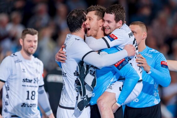Eine Gruppe männlicher Handballspieler feiert fröhlich auf dem Spielfeld, wobei ein Spieler in einem blauen Trikot einem Mannschaftskameraden in einem weißen Trikot in die Arme springt. Im Hintergrund ist eine unscharfe Menschenmenge zu sehen.