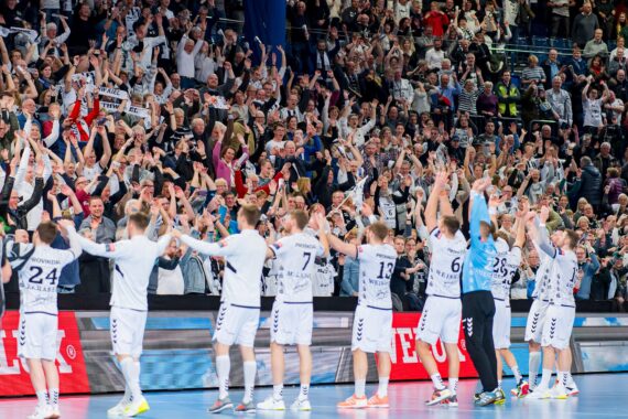 Eine Männerhandballmannschaft in weißen Trikots steht in einer Reihe auf dem Spielfeld und hebt die Arme, um mit einer großen, jubelnden Menge in der Arena hinter ihnen zu feiern.