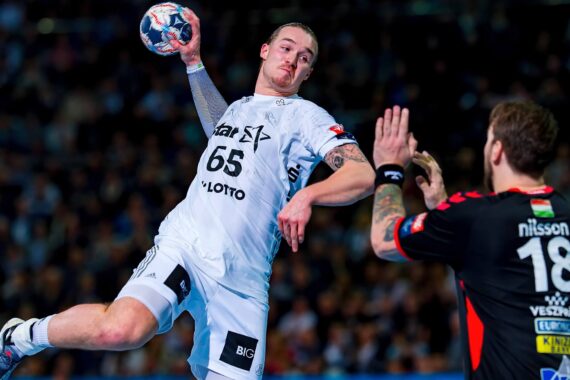 Ein männlicher Handballspieler in einem weißen Trikot bereitet sich in der Luft darauf vor, den Ball zu werfen, während ein anderer Spieler in einem schwarzen Trikot mit ausgestreckten Armen versucht, ihn in einer überfüllten Halle zu blockieren.