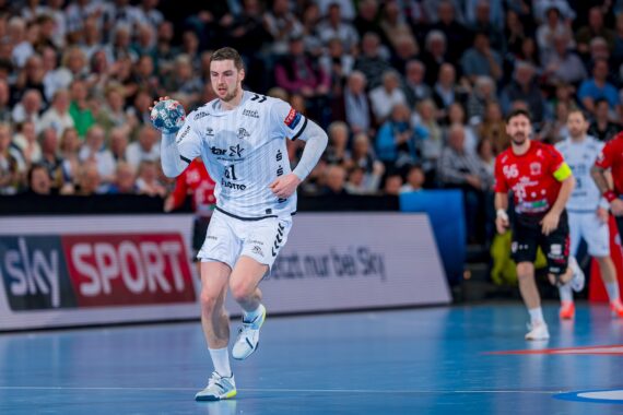 Ein männlicher Handballspieler in einem weißen Trikot läuft während eines Spiels mit dem Ball, während die gegnerischen Spieler in roten Trikots und eine große Menschenmenge im Hintergrund zusehen.