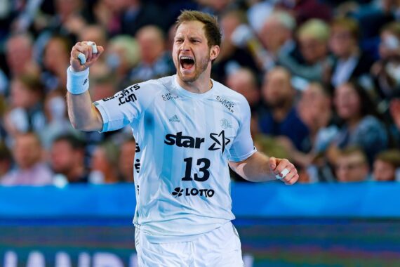 Ein männlicher Handballspieler, der ein weißes Trikot mit der Nummer 13 trägt, jubelt während eines Spiels mit einer geballten Faust, umgeben von einer unscharfen Menschenmenge im Hintergrund.