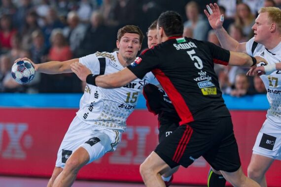 Ein Handballspieler in einem weißen Trikot bereitet sich darauf vor, den Ball zu werfen, während er während eines Spiels von Gegnern in schwarzen Trikots eng bewacht wird. Die Aktion findet auf einem Spielfeld mit einer Menschenmenge im Hintergrund statt.