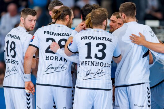 Eine Gruppe männlicher Handballspieler in weißen Trikots mit der Aufschrift PROVINZIAL auf dem Rücken steht in einem Pulk, von der Kamera abgewandt, und bereitet sich auf ein Spiel vor oder bespricht eine Strategie.