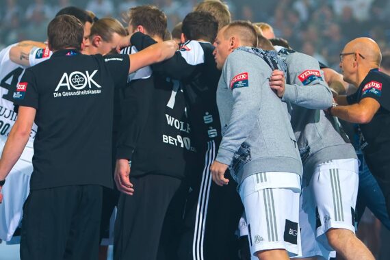 Eine Gruppe männlicher Athleten in Sportuniformen versammelt sich in einem Mannschaftskreis und wirkt vor einem Spiel oder Wettkampf konzentriert und hilfsbereit. Trainer und Betreuer stehen in der Nähe, und im Hintergrund ist eine Menschenmenge zu sehen.