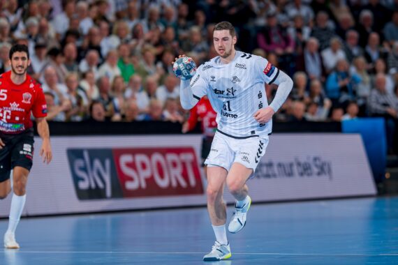 Ein männlicher Handballspieler in einem weißen Trikot läuft während eines Spiels mit dem Ball, während die Gegner in roten Trikots ihn auf einem Hallenplatz verfolgen. Im Hintergrund ist eine Menschenmenge zu sehen, die das Spiel verfolgt.