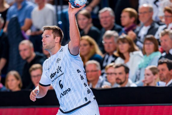 Ein männlicher Handballspieler in einem weißen Trikot mit der Nummer 6 bereitet sich während eines Spiels auf den Wurf vor, während im Hintergrund eine Menge von Zuschauern zusieht.